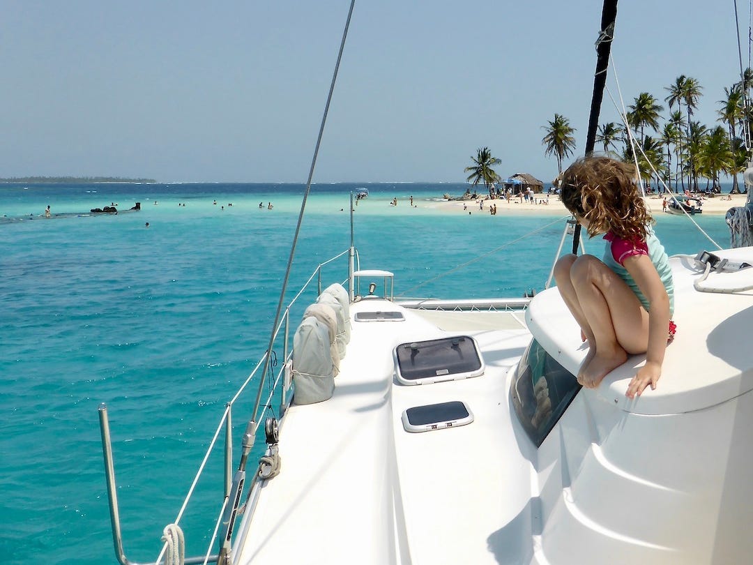 A young girl sits on top of a white yacht on the left surrounded by water with an island in the background